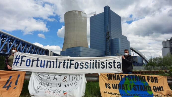 Kaksi ihmistä pitelee banderollia, jossa lukee #FortumIrtiFossiilisista. Taustalla hiilivoimala.