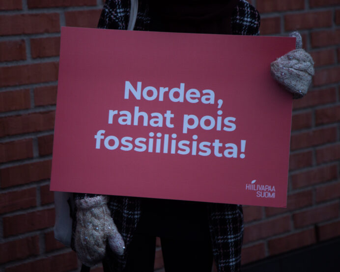 Kädet pitelevät punaista mielenosoituskylttiä, jossa lukee Nordea, rahat pois fossiilisista.