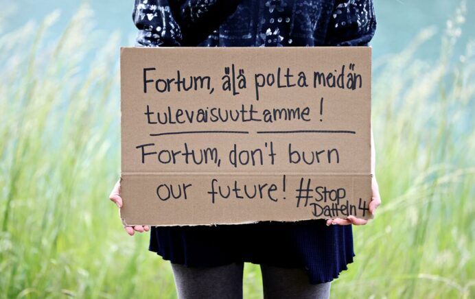 Mustaan pukeutunut henkilö sinivihreän taustan edessä pitää kylttiä, jossa lukee: Fortum älä polta meidän tulevaisuuttamme! Fortum don't burn our future! #StopDatteln4